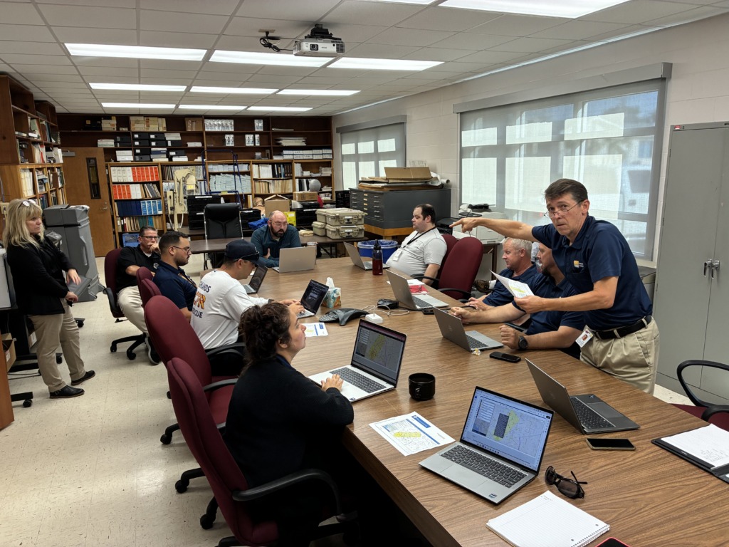 A group of people in a conference room, seated around a large table with laptops and maps. One man stands, pointing and holding a map, while others are engaged with their screens.