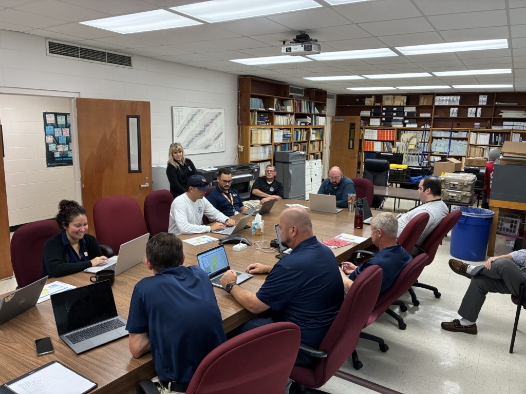 A group of ten people, mostly men, are seated around a large conference table in an office, working on laptops. One woman stands at the back, and another woman smiles while seated at the table. Bookshelves and posters are in the background.