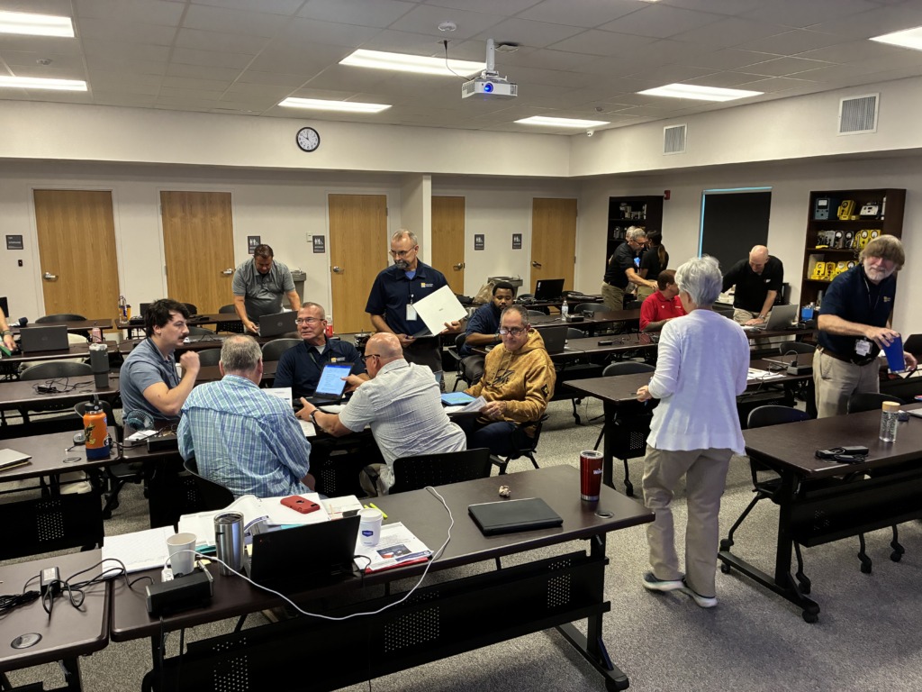 A group of adults, mostly men, in a brightly lit classroom. Many are seated at individual desks with laptops, while others stand and interact, suggesting a collaborative training session.