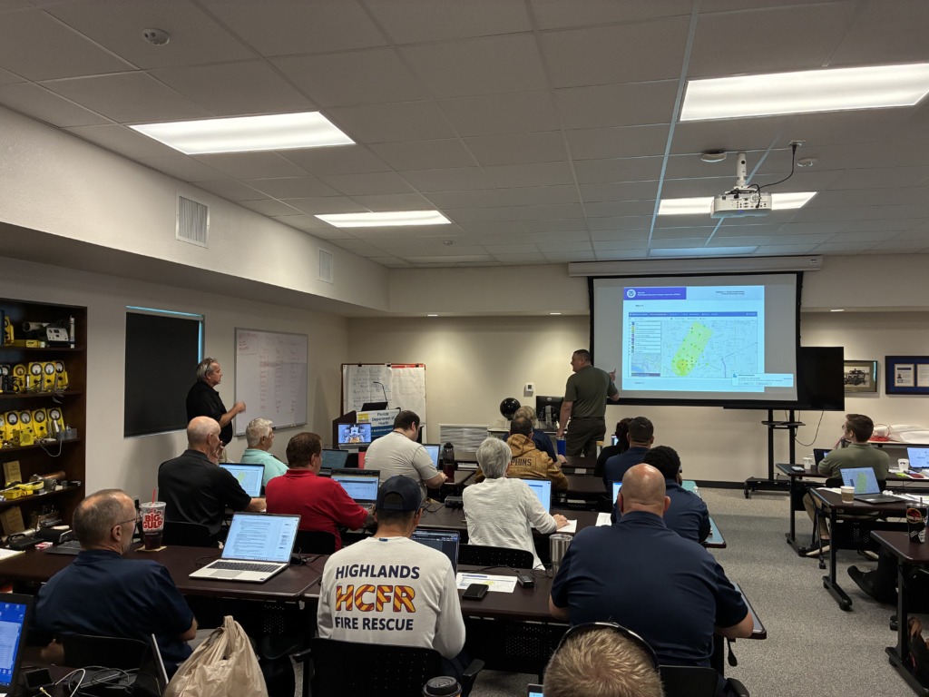 A group of adults, many with laptops, attending a training session in a classroom. A projector screen displays a map, and an instructor gestures towards it. Shelves with technical equipment are visible on the left.