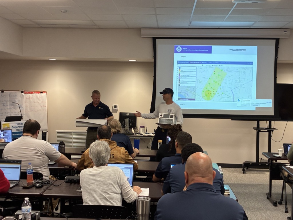 A group of people attending a training session on radiological operations. Two instructors are at the front, one holding a laptop and gesturing towards a large screen displaying a map with highlighted contamination zones, while the other holds a rolled-up map. Participants are seated at tables with laptops.