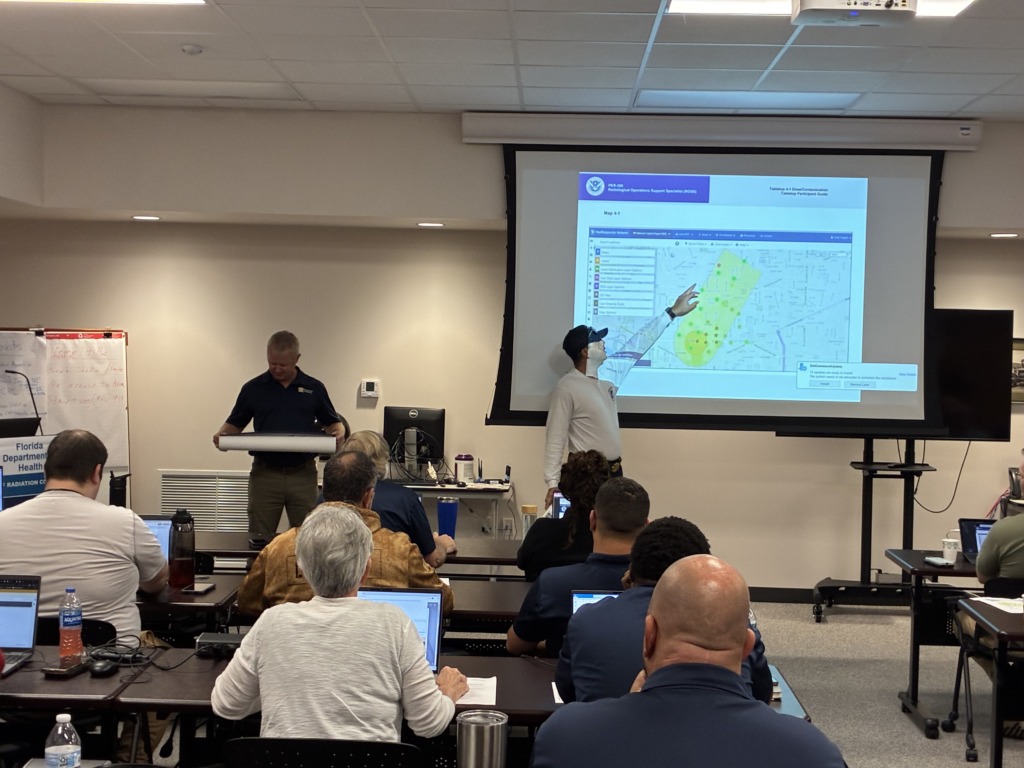 A group of people attends a radiological operations training session. Two instructors are at the front, one pointing at a large screen displaying a map with highlighted areas, while the other holds a rolled-up document. Attendees are seated at tables with laptops.