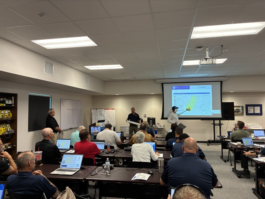 A group of adults attends a training session in a classroom, with a presenter pointing at a projected map on a screen and attendees using laptops.