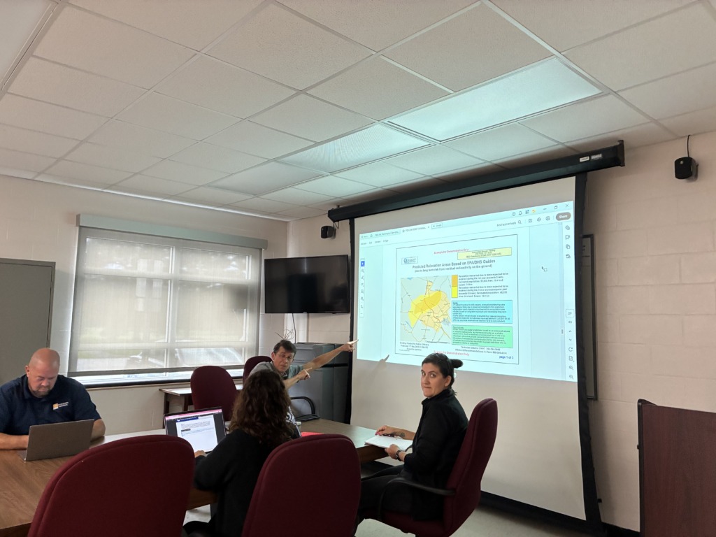 Four people in a meeting room. A man points at a large projector screen displaying a map of predicted relocation areas and related data, while others are seated around a table with laptops and notebooks.
