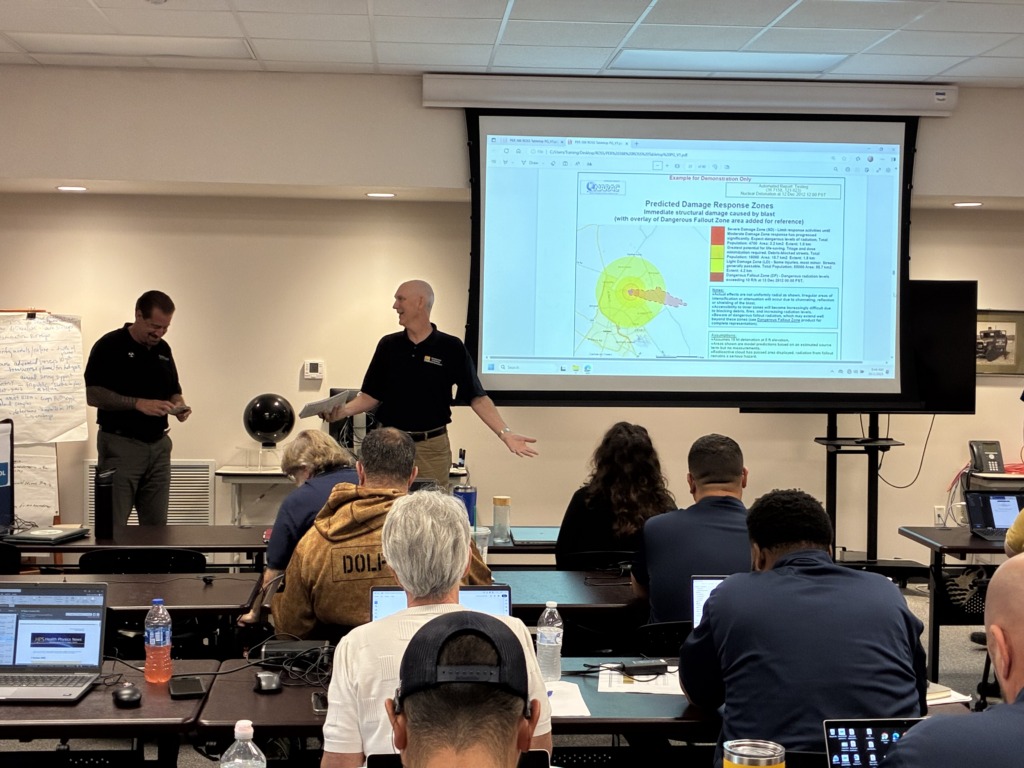 A presenter gestures towards a projector screen showing a map of predicted damage and fallout zones from a simulated blast, while attendees sit at tables with laptops during a training session.