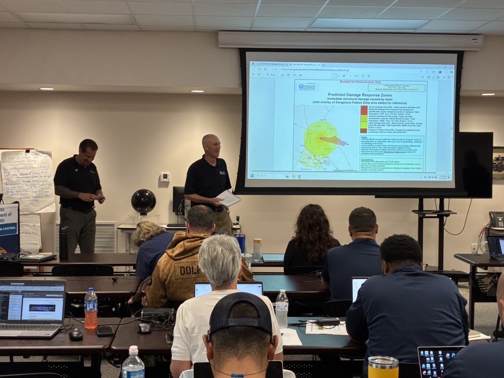 A group of people attending an emergency response training, with a presenter in front of a screen displaying a map of predicted blast damage and fallout zones from a simulated nuclear detonation.