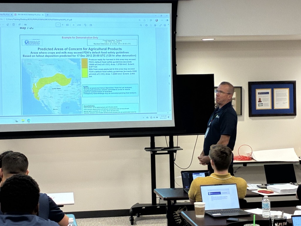 A man presents a slide showing a map of the United States with yellow-shaded areas indicating predicted agricultural contamination zones, to an audience in a classroom setting.