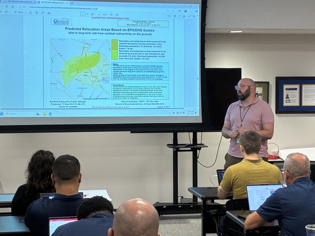 A man with a beard and glasses presents to an audience in a classroom, standing next to a large screen displaying a map and text about predicted relocation areas due to residual radioactivity based on EPA/DHS guides.