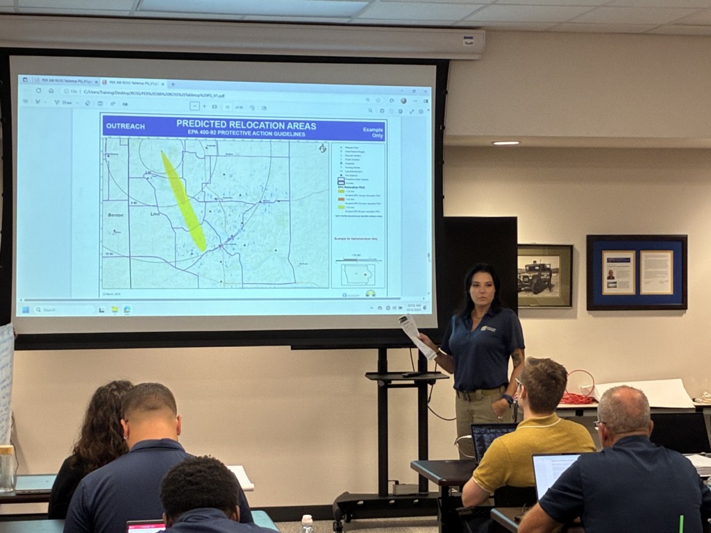 A woman presents a map of predicted relocation areas and EPA guidelines on a projector screen to a group of seated attendees in a training room.