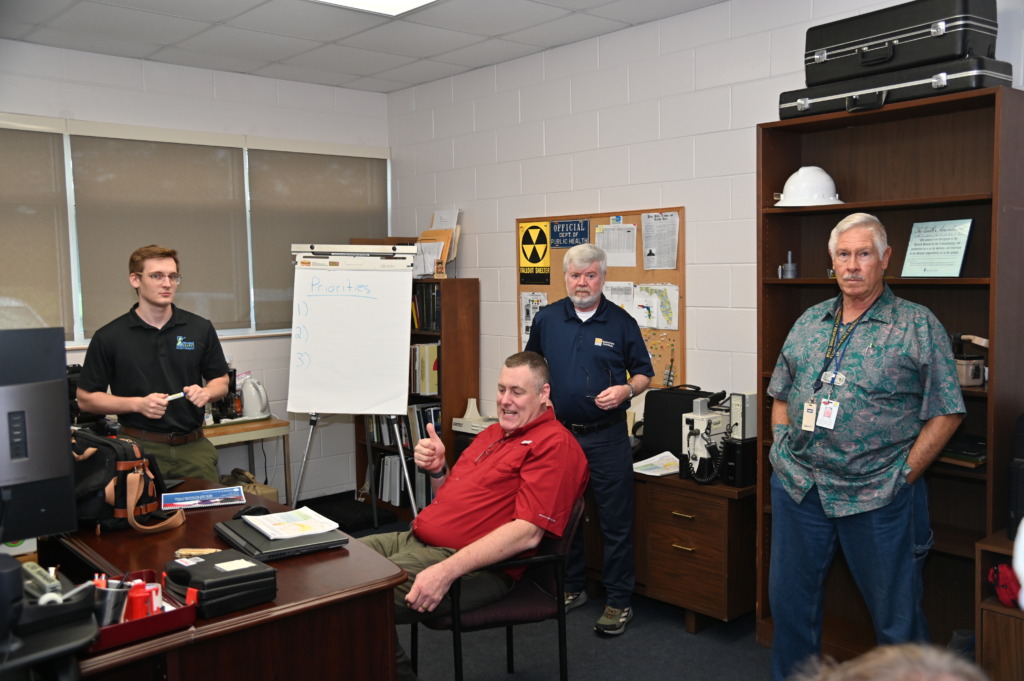 Four men in an office setting, one seated giving a thumbs-up, others standing near a whiteboard listing 'Priorities' and a bulletin board with 'Fallout Shelter' and 'Public Health' signs.
