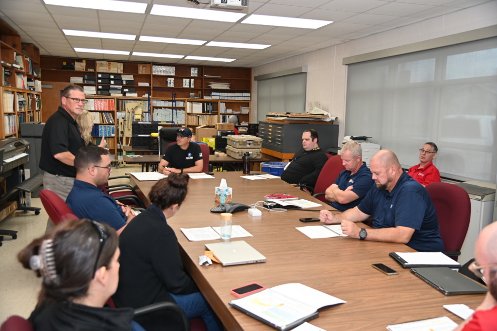 A group of about ten people, mostly men, are seated and standing around a large wooden conference table in a room filled with shelves of binders, engaged in a meeting.