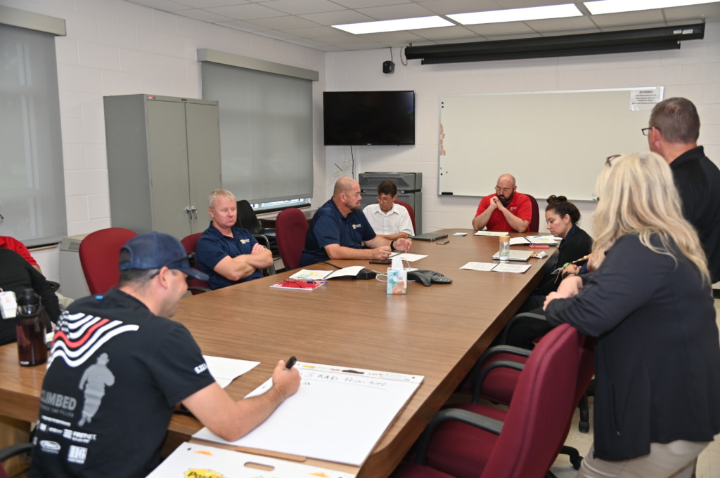 A group of people attending a meeting around a large wooden conference table in a room with white walls and a whiteboard.