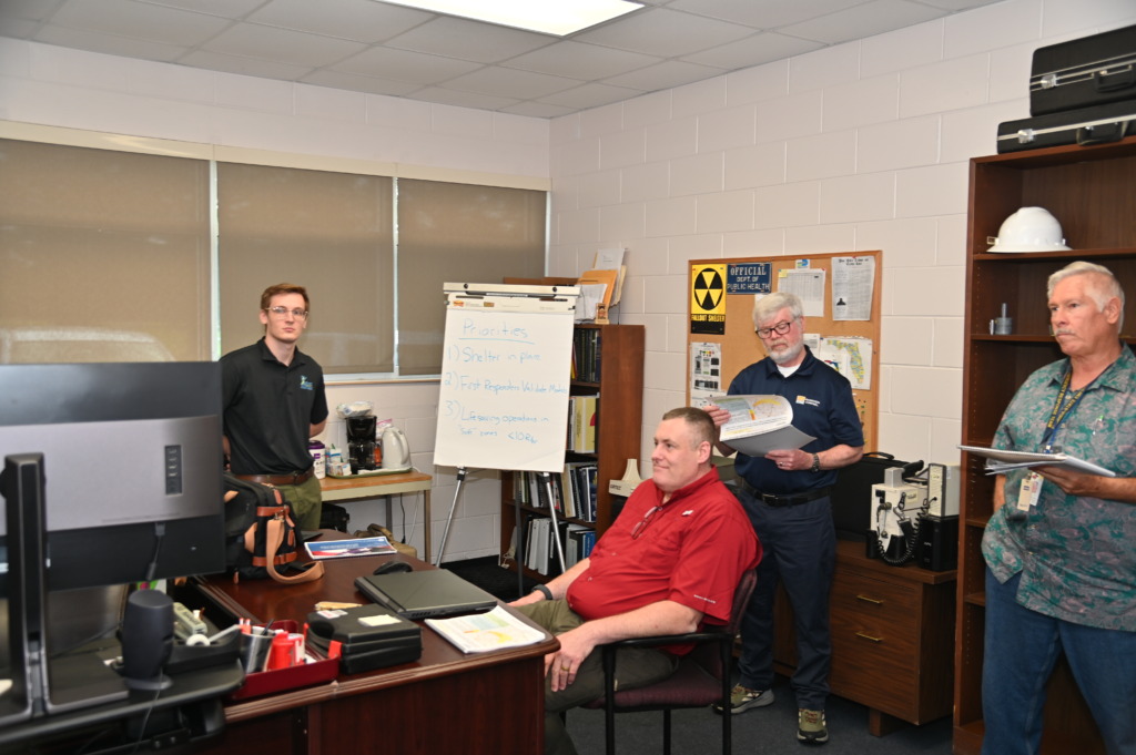 Four men in an office setting discussing emergency preparedness. A whiteboard lists priorities like 'Shelter in place' and 'Lifesaving operations.' Signs for 'Fallout Shelter' and 'Dept. of Public Health' are visible on a corkboard.
