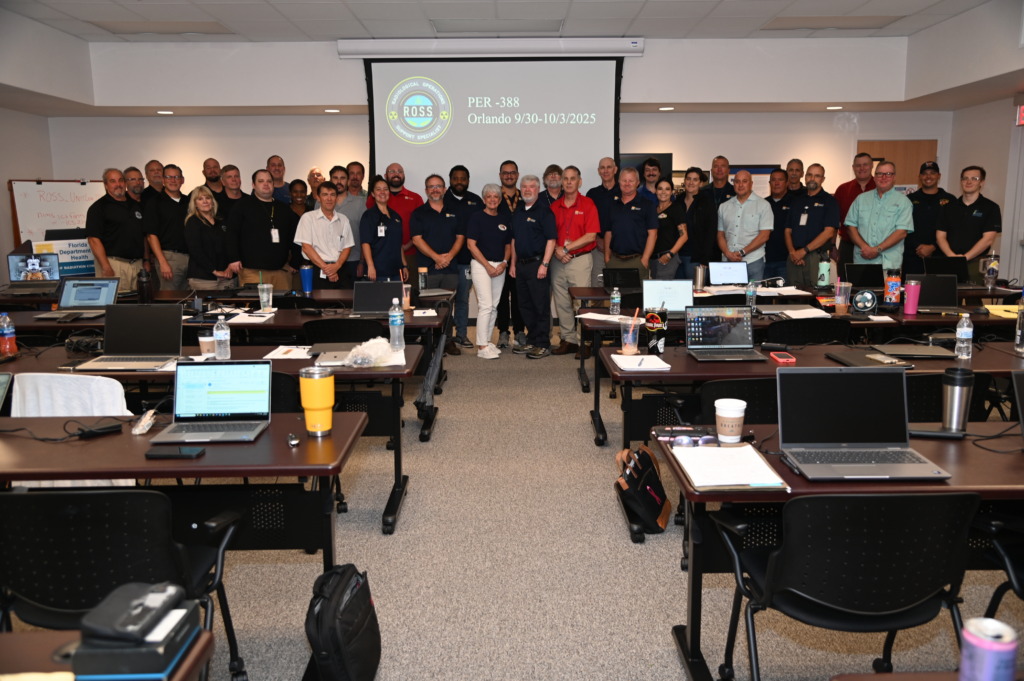 A large group of about 35-40 adults, many in polo shirts, stand smiling in a training room with laptops on tables, in front of a screen displaying 'R.O.S.S.' and 'Orlando 9/30-10/3/2025'.