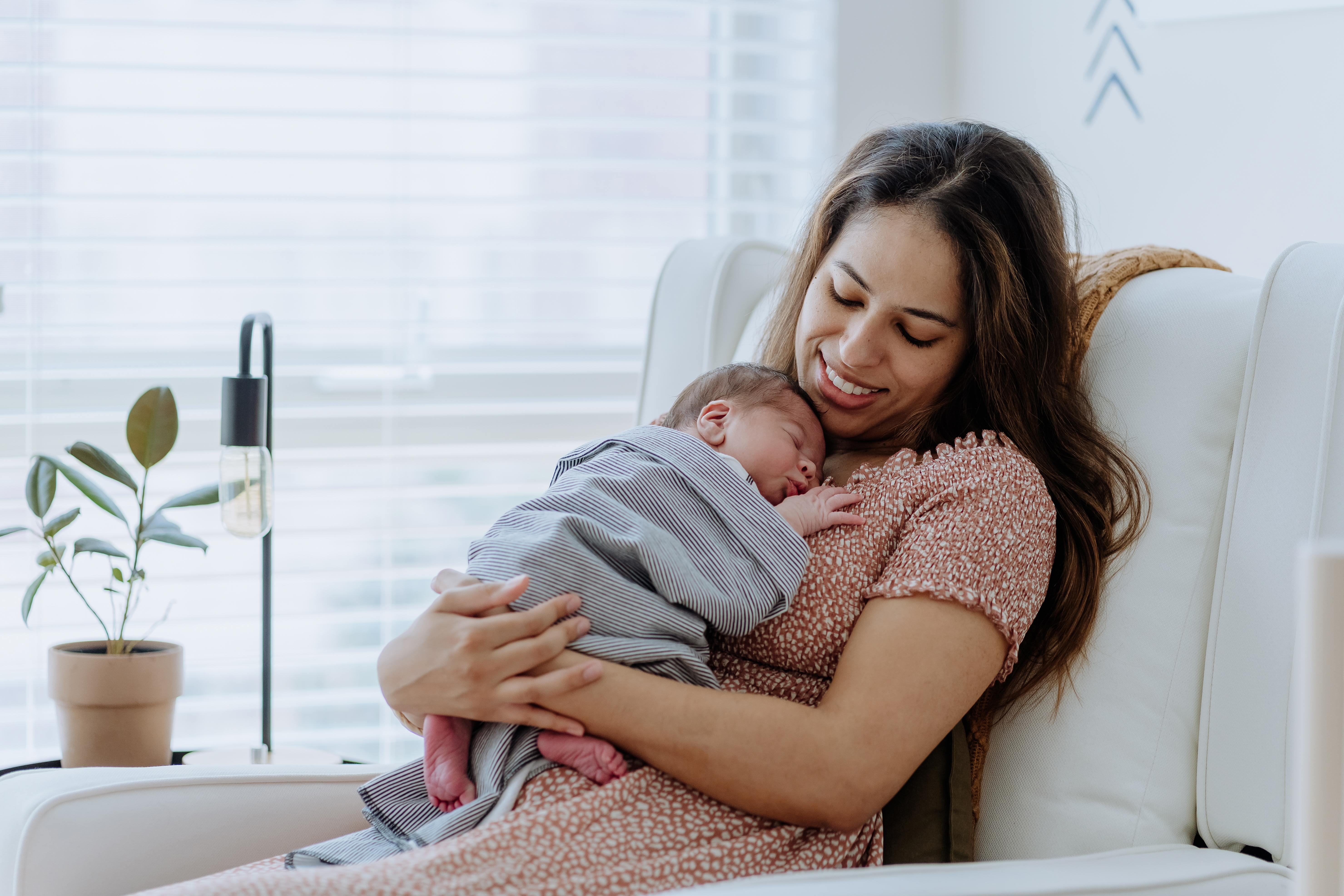 A smiling mother with long dark hair holds her newborn baby, who is swaddled in a striped blanket and sleeping peacefully against her chest. They are seated in a white armchair by a window with blinds.