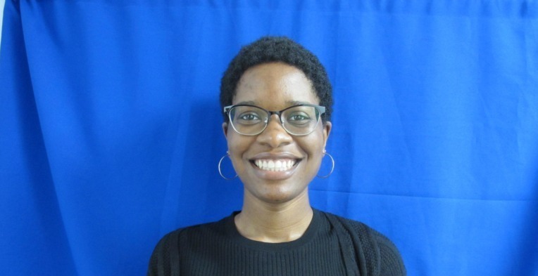 Headshot of a smiling Black woman with short curly hair and glasses, wearing a black top, against a blue background.