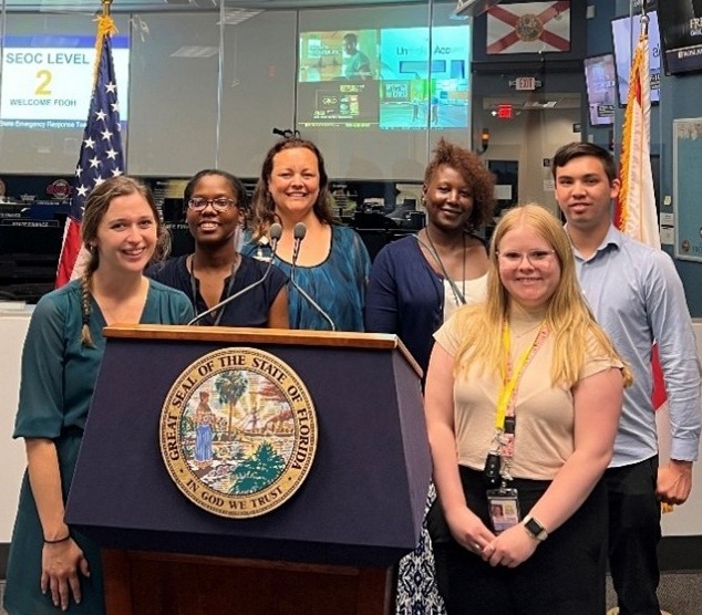 Six people, four women and two men, smiling behind a podium with the Florida State Seal, in front of an American flag and a Florida flag, inside an emergency operations center with screens displaying 'SEOC LEVEL 2'.
