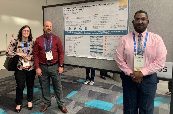 Three people, two men and one woman, stand smiling in front of a large research poster board at a conference.