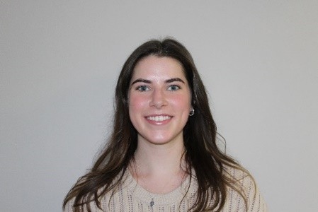 Headshot of a smiling young woman with long brown hair and a light-colored sweater against a plain background.