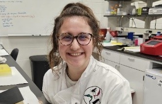 A smiling young woman in a lab coat and glasses, wearing black gloves, stands in a lab with a large plume of white vapor rising from a container in front of her.