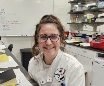A smiling young woman in a white lab coat and glasses works with a container emitting a large plume of white vapor on a lab bench.