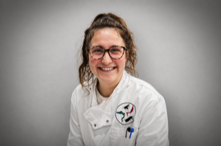 A smiling woman with light brown hair and glasses, wearing a white chef's coat with a circular logo patch.