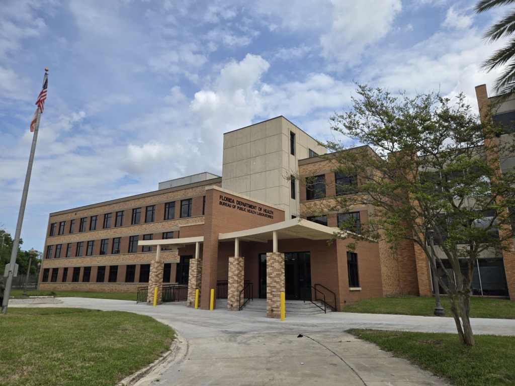 Exterior view of the Florida Department of Health building, a multi-story brick and concrete structure, with an American flag flying on a flagpole and green lawns.