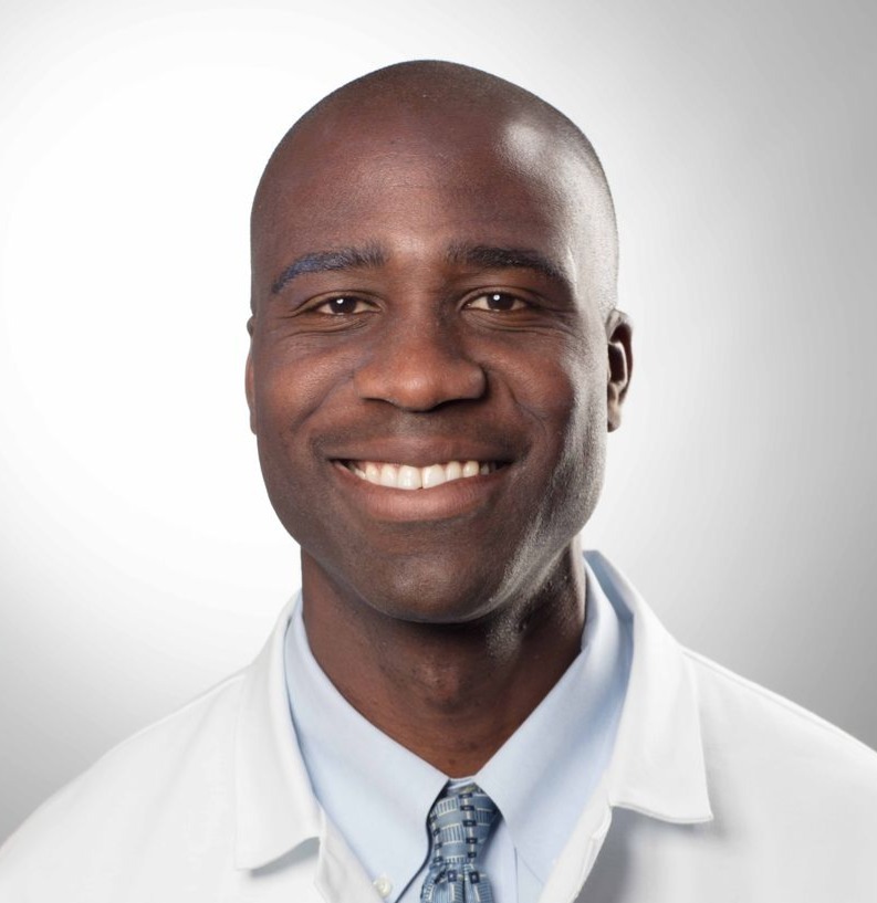 Headshot of a smiling bald man in a white lab coat, blue shirt, and patterned tie.