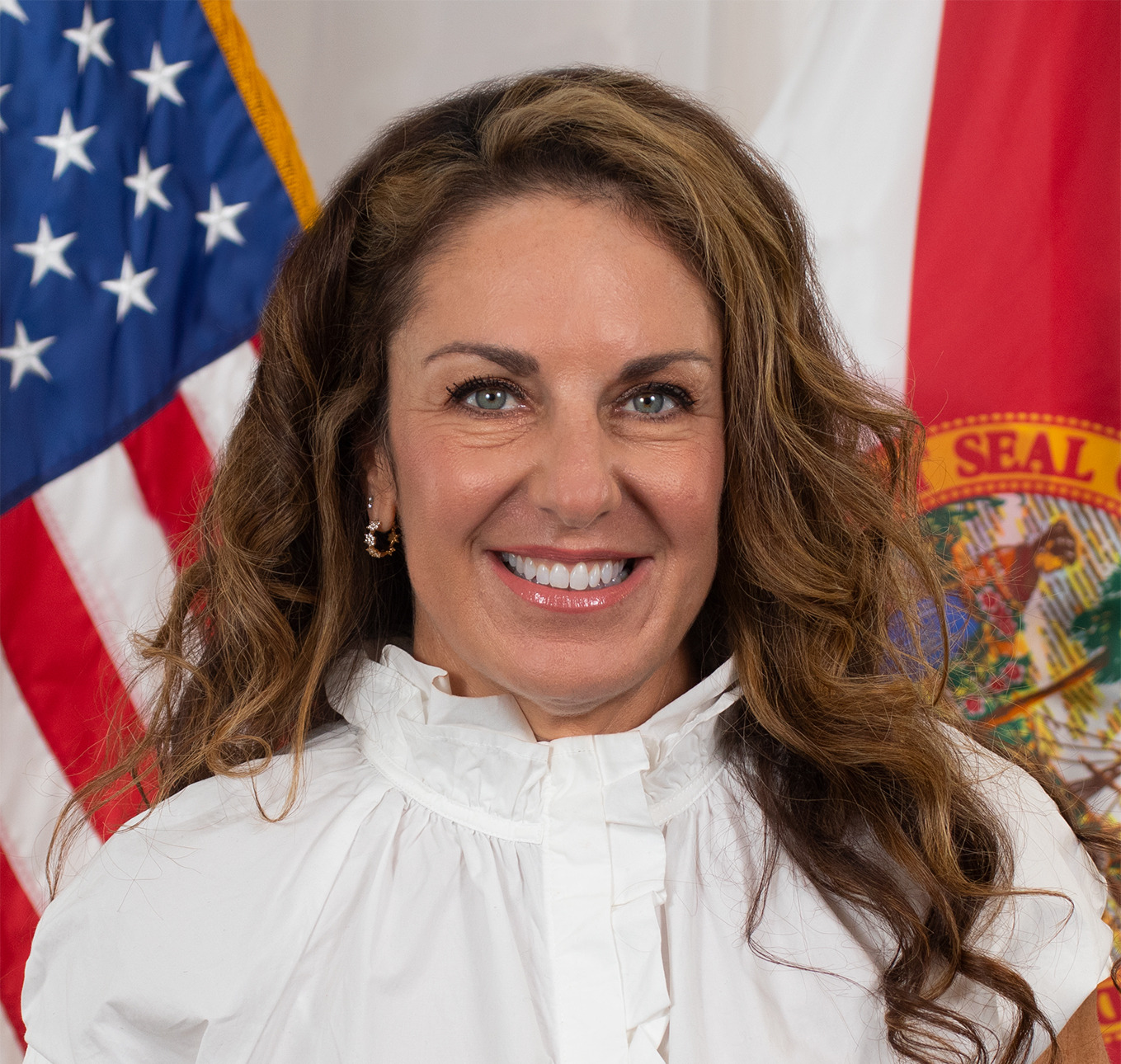 A smiling woman with brown hair and blue-green eyes in a white ruffled blouse, flanked by the American and Florida flags.