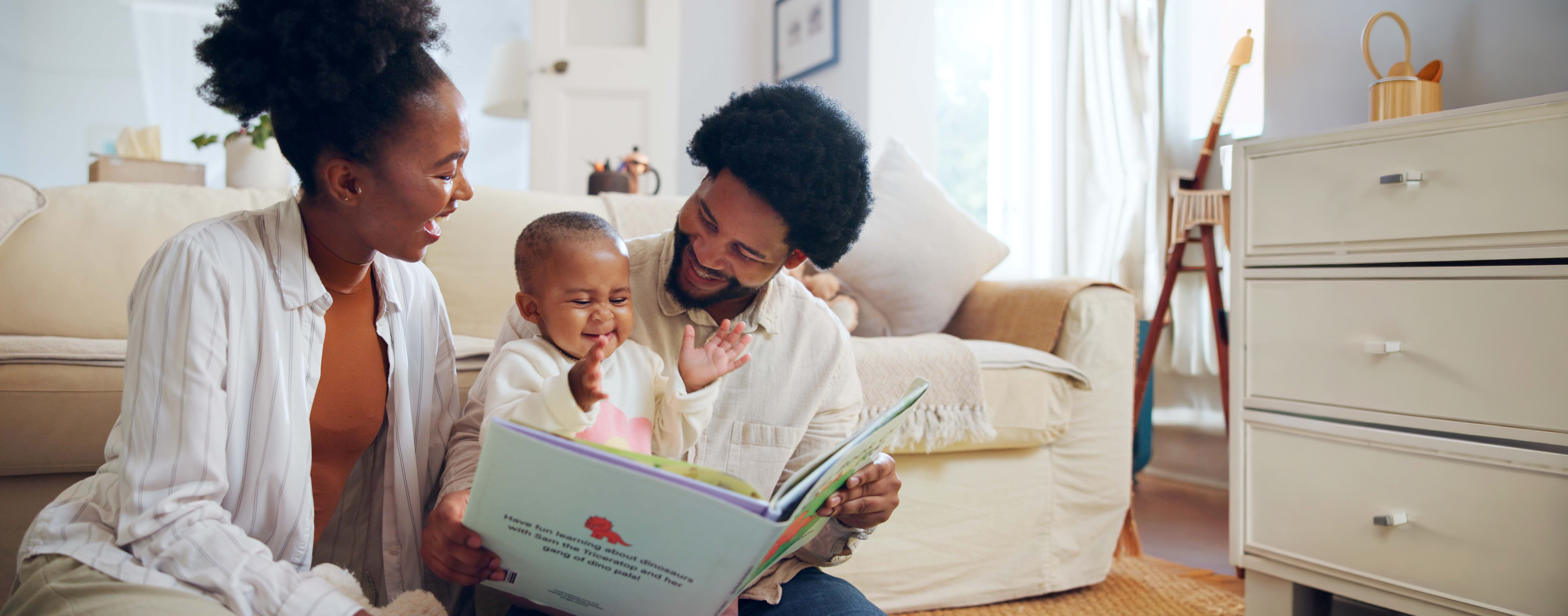 Parents smiling at their baby, who is laughing with hands up, while reading a dinosaur book together on the floor.