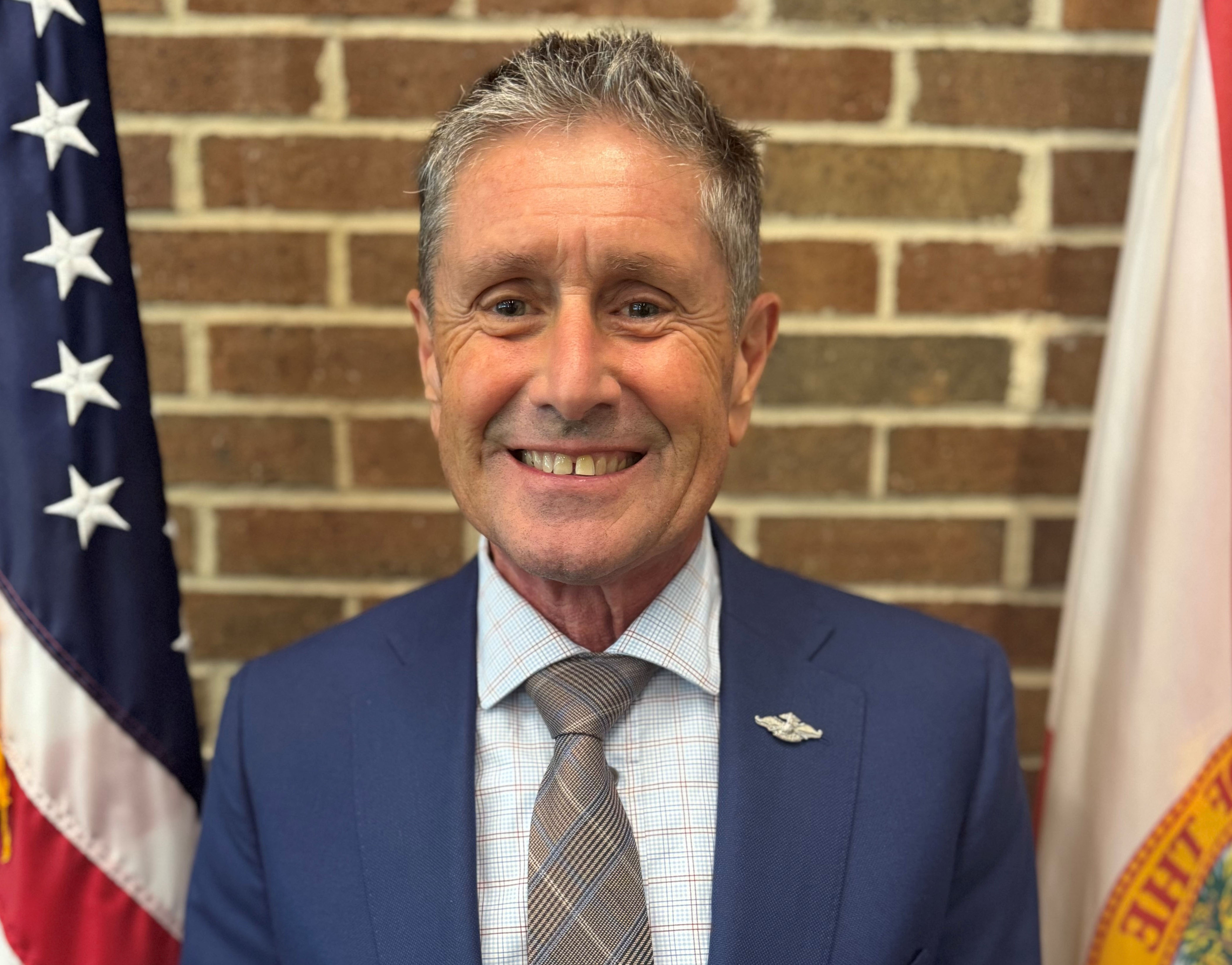 Portrait of a smiling man in a blue suit and plaid tie, with the American flag on his left and the Florida state flag on his right, against a brick wall.