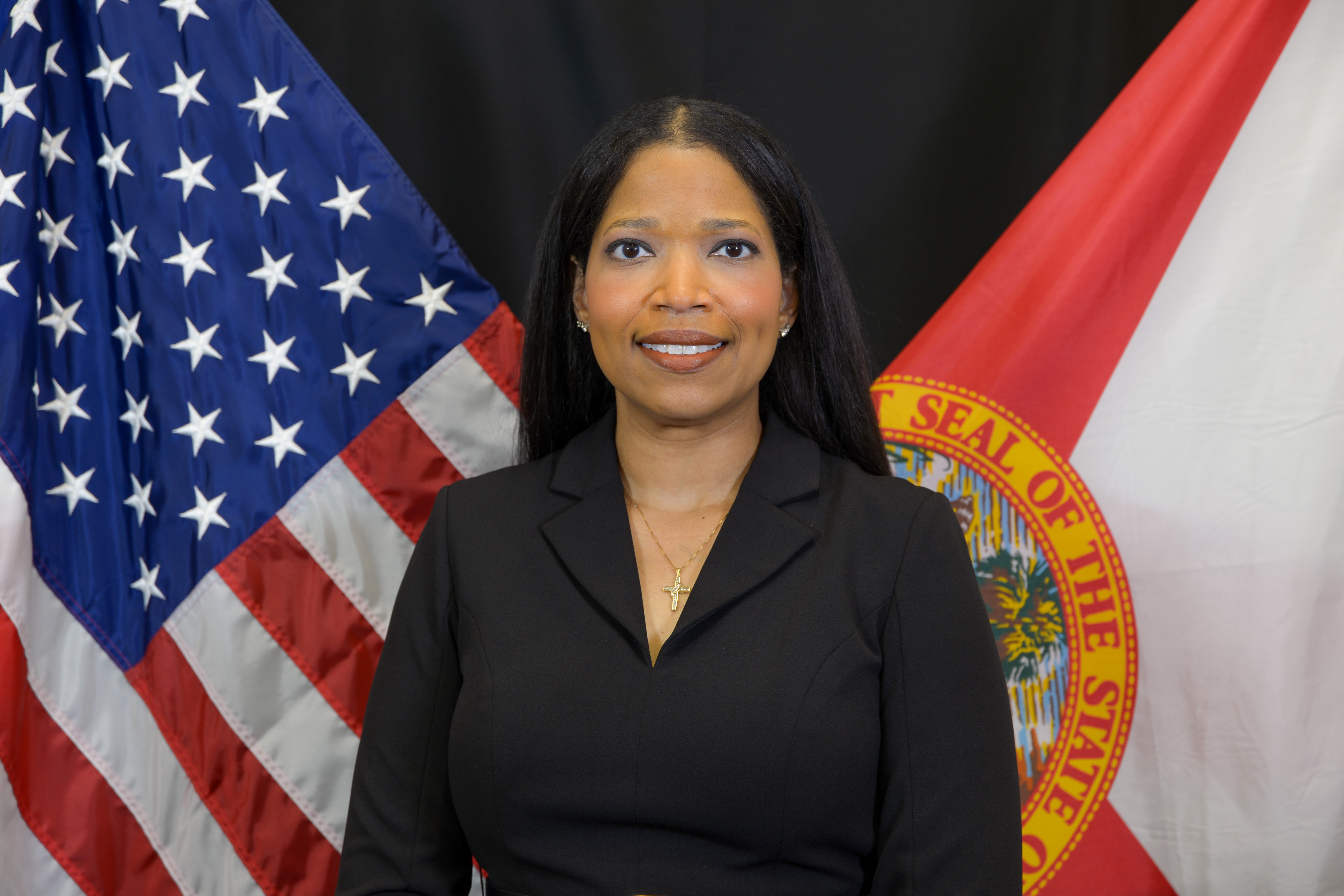 A woman in a black blazer smiles, with the American flag on her left and the Florida state flag on her right.