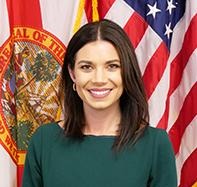 Headshot of a smiling woman in a dark green dress, with the Florida state flag and US flag in the background.