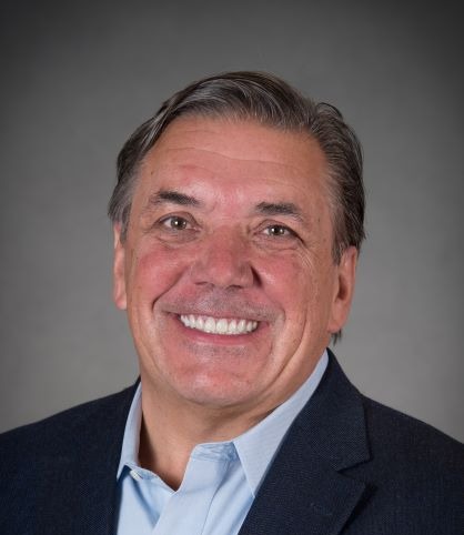 Headshot of a smiling man with graying hair, wearing a light blue shirt and dark blazer.