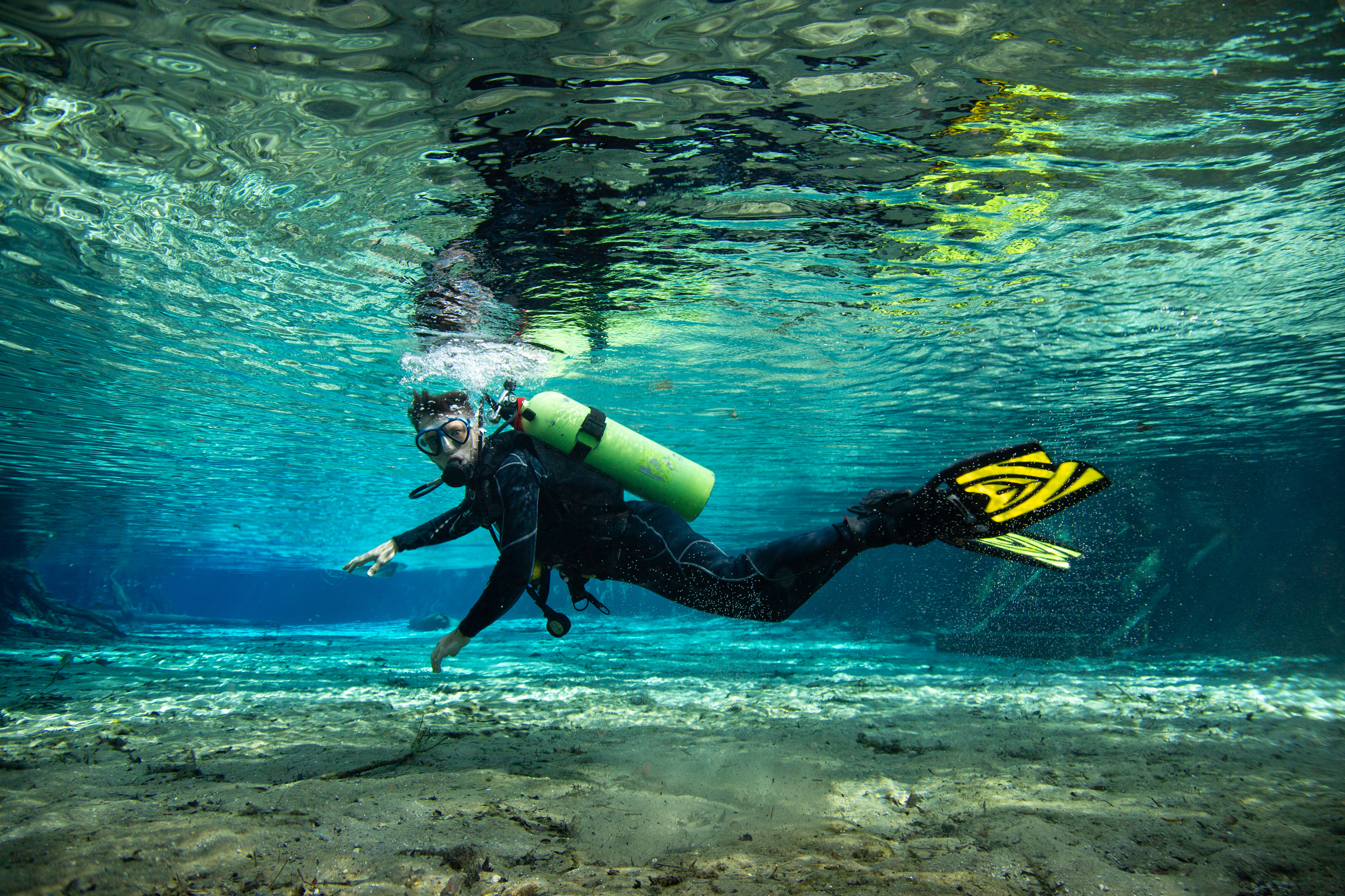 A scuba diver in a black wetsuit and yellow-green tank swims horizontally in clear, turquoise water, with a sandy bottom visible below and rippling surface above.