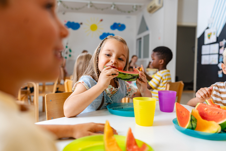 A young girl eats a large slice of watermelon, while sitting at a table with other children in a colorful classroom.