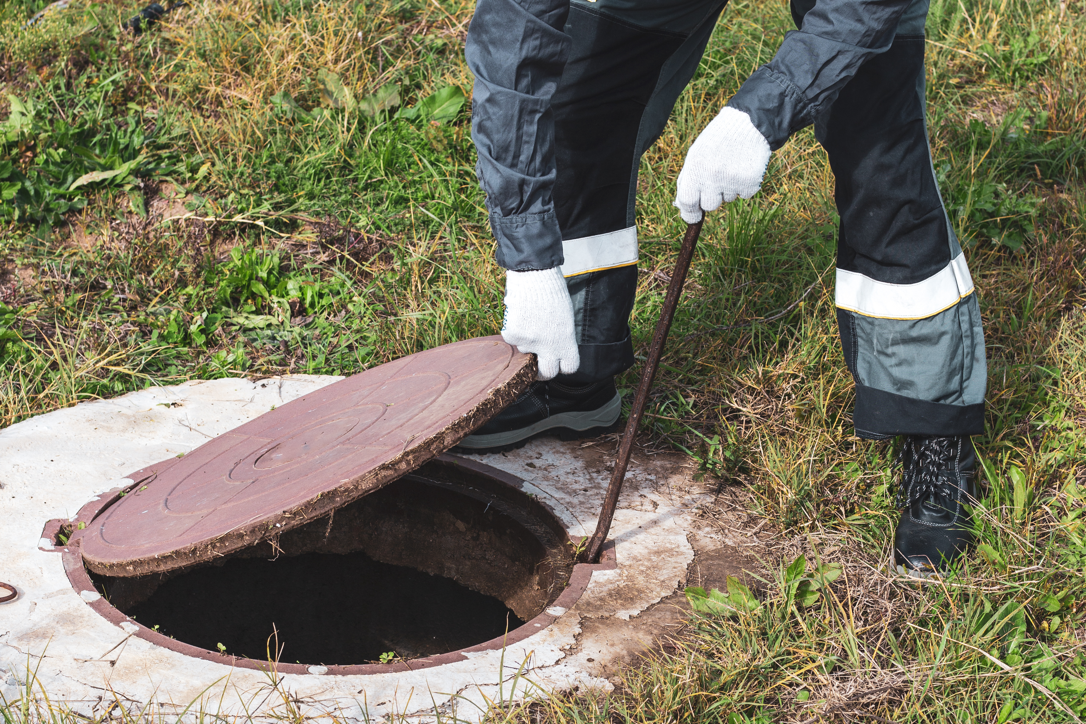 A person in work pants and white gloves uses a metal bar to open a manhole cover